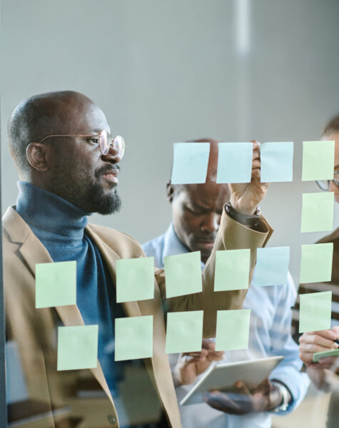 Young confident businessman in casualwear writing down tasks on notepapers while standing in front of transparent board